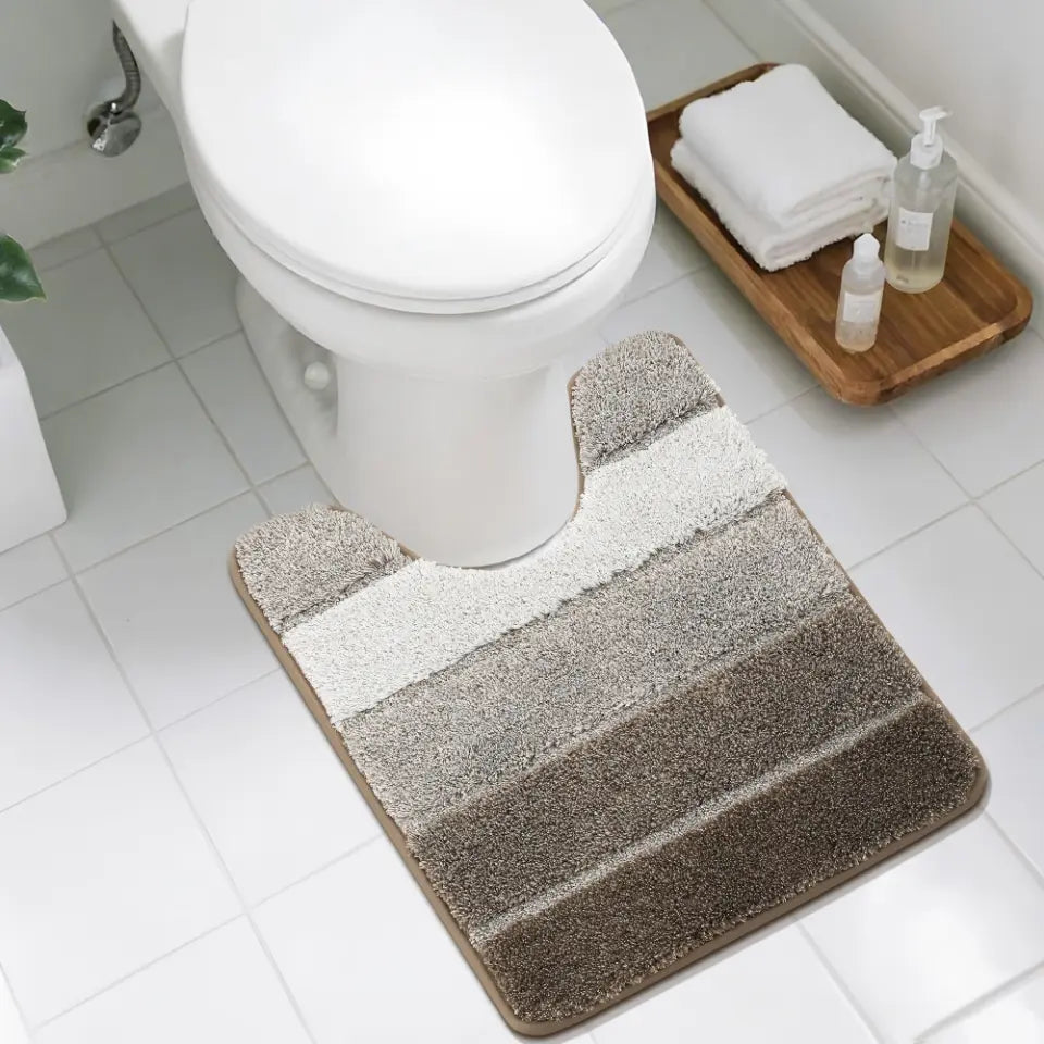Bathroom with a brown striped U shaped bath mat in front of a toilet, a wooden tray with towels and bottles in the background.