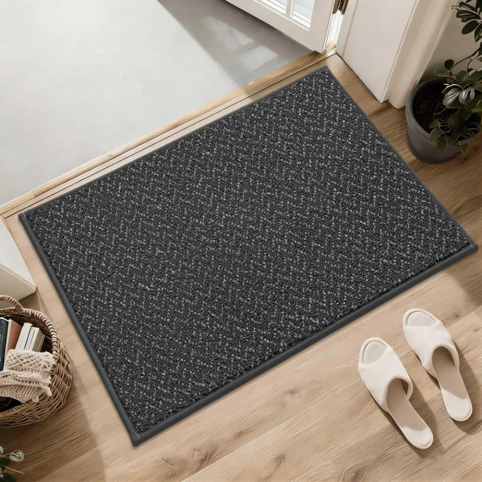 Dark grey doormat on a wooden floor with white slippers and a plant in the background.